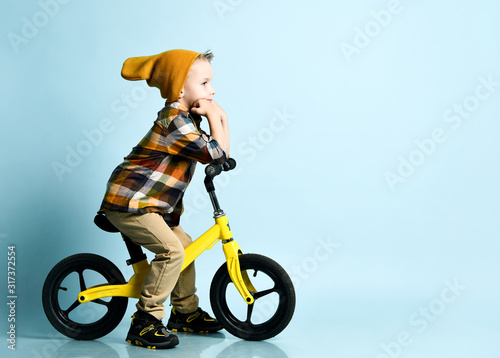 Positive boy in bright stylish casual clothing, sneakers and cap sitting on yellow bibycle present over blue background photo