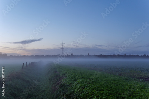 Beautiful dramatic twilight gold and blue cloud and sky over misty atmosphere of foggy over agricultural field and high voltage tower on countryside in Germany during sunset time.