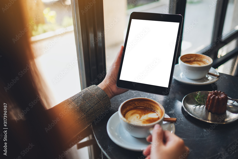 Mockup image of a woman sitting and holding black tablet pc with blank white desktop screen while drinking coffee in cafe - obrazy, fototapety, plakaty 