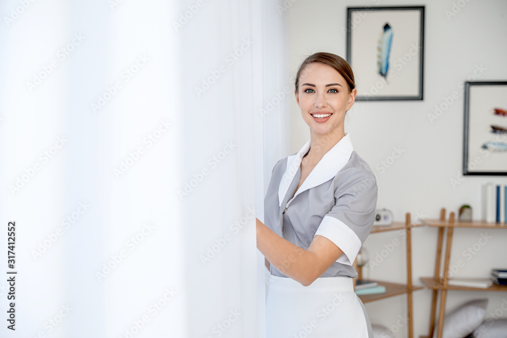Beautiful young chambermaid opening curtains in hotel room Beautiful young chambermaid opening curtains in hotel room