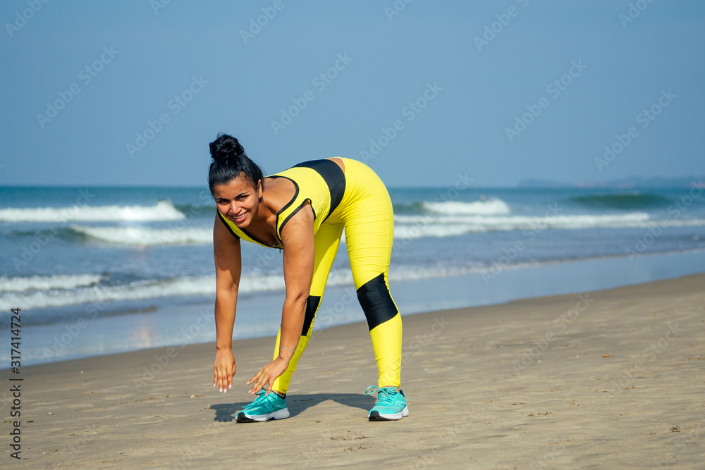 happy asian brunette woman in stylish yellow leggings and fitness bra training body on the beach in morning Goa