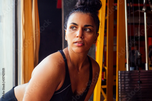 latin american fitness model in bra top and leggings looking tired with sweat on the forehead at dark gym