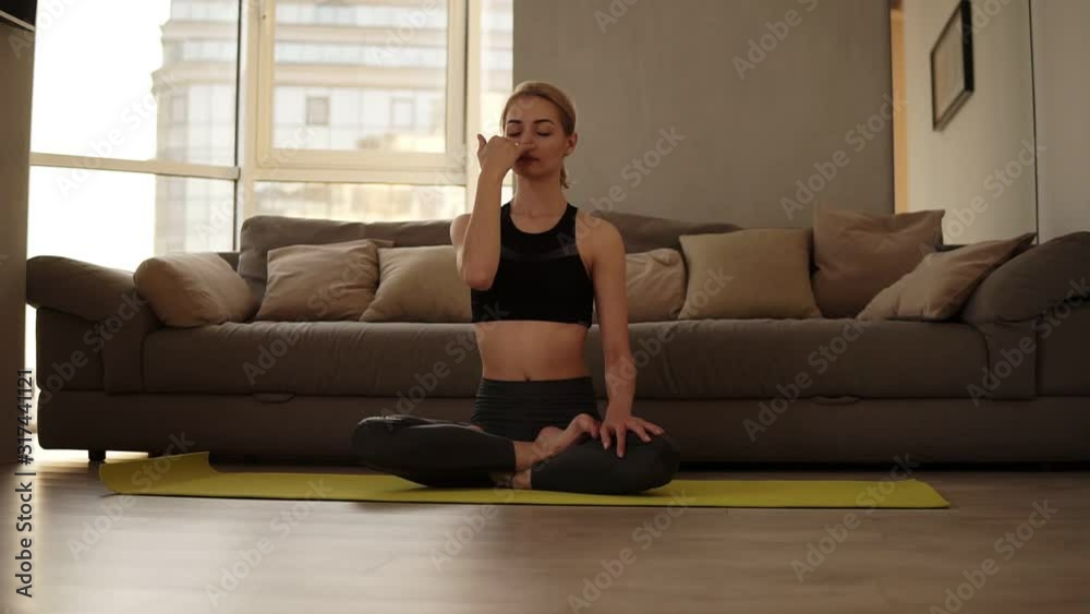 Young attractive woman practicing yoga exercise, sitting in lotus pose, performing Alternate Nostril Breathing technique indoors. Making nadi shodhana pranayama therapeutic exercise Young attractive woman practicing yoga exercise, sitting in lotus pose, performing Alternate Nostril Breathing technique indoors. Making nadi shodhana pranayama therapeutic exercise