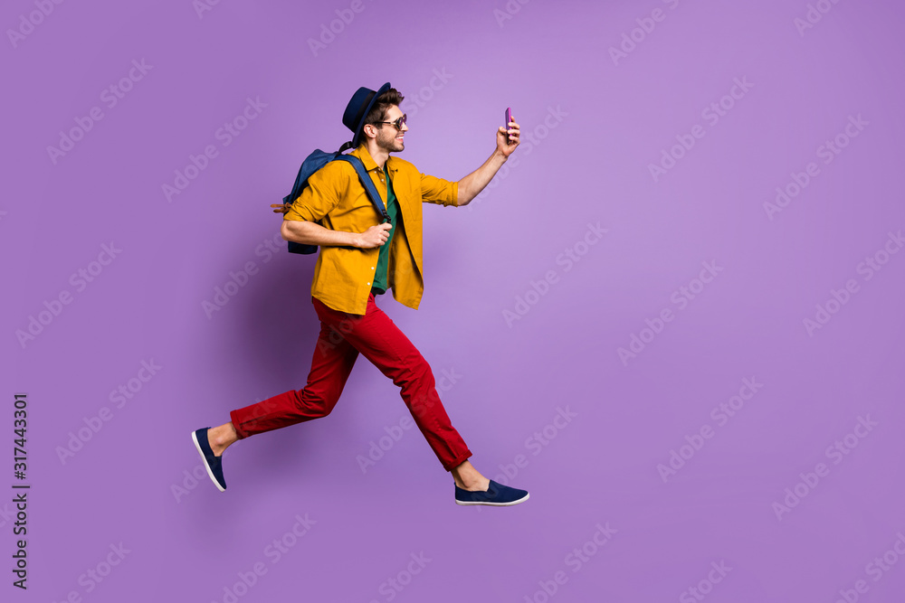 Full size profile side photo of cheerful guy in retro headwear holding blue bag jump run make selfie on smartphone wear yellow shirt red pants trousers isolated over violet color background