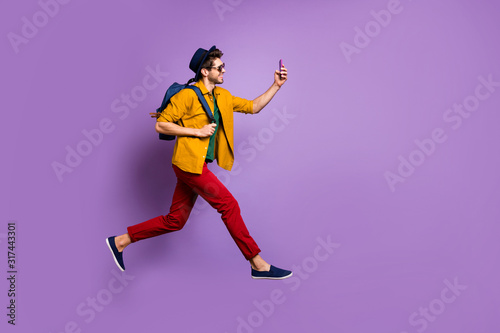 Full size profile side photo of cheerful guy in retro headwear holding blue bag jump run make selfie on smartphone wear yellow shirt red pants trousers isolated over violet color background