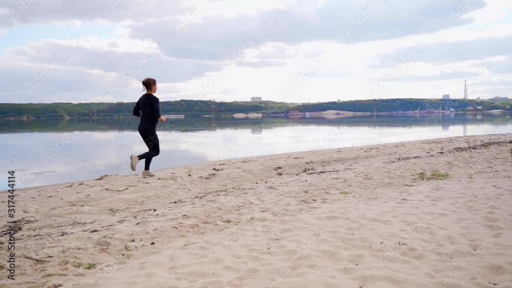 A girl in black clothes and headphones runs along the sandy bank of the river. A girl in black clothes and headphones runs along the sandy bank of the river.