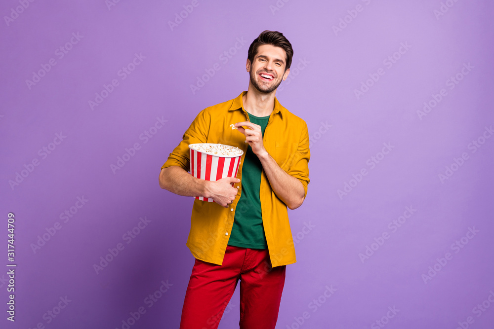 Portrait of positive cheerful guy have free time hold big pop corn box watch tv funny series laugh wear youth modern clothes isolated over violet color background Portrait of positive cheerful guy have free time hold big pop corn box watch tv funny series laugh wear youth modern clothes isolated over violet color background