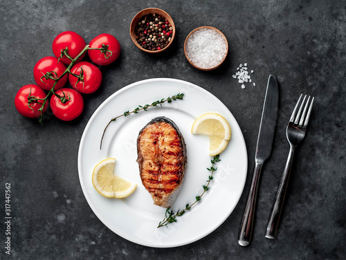 Grilled salmon steak in a white plate with spices, fork and knife on a stone background