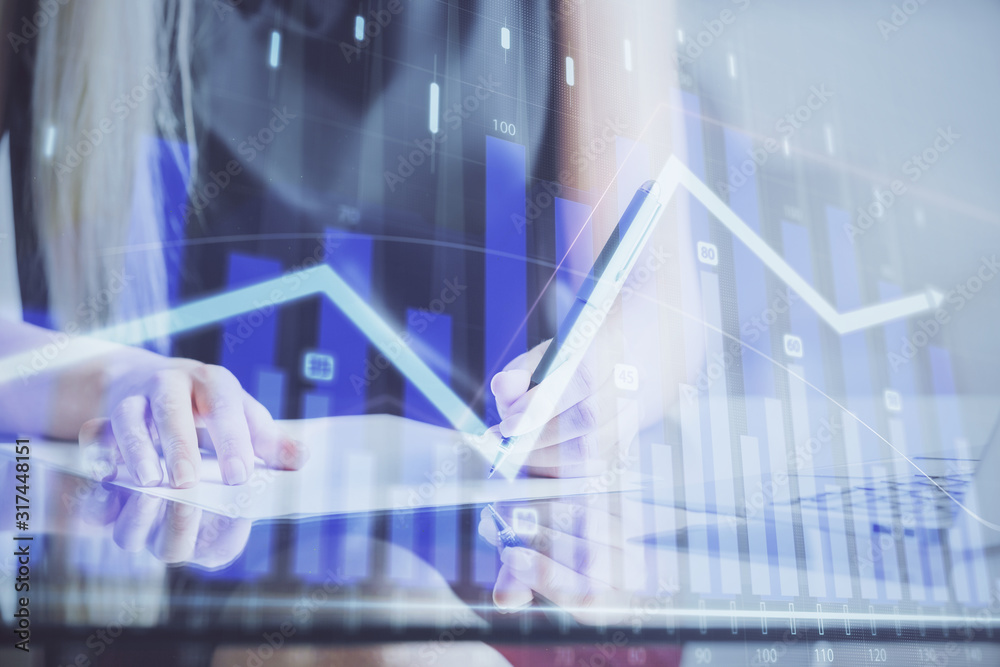 A woman hands writing information about stock market in notepad. Forex chart holograms in front. Concept of research. A woman hands writing information about stock market in notepad. Forex chart holograms in front. Concept of research.