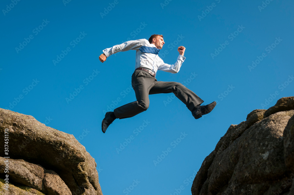 Courageous businessman jumping between two boulders outdoors above a steep crevasse against bright blue sky Courageous businessman jumping between two boulders outdoors above a steep crevasse against bright blue sky