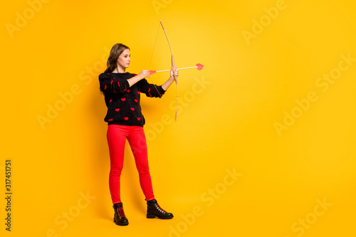 Full length profile photo of pretty lady working as cupid hold bow arrow aiming feelings love couple wear hearts pattern sweater red trousers shoes isolated yellow color background