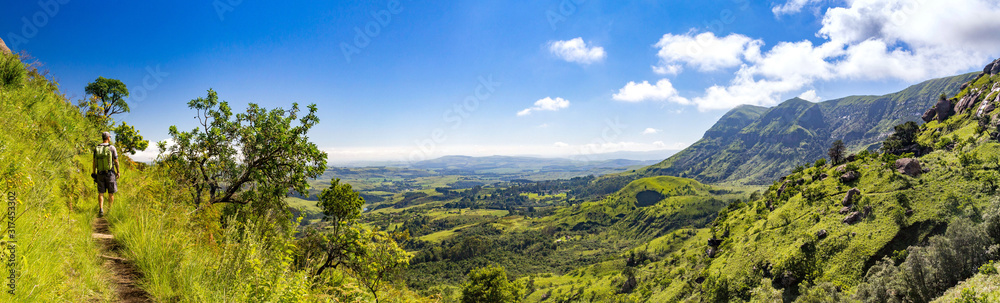 Man hiking on a small trail with a panoramic view over steep and green mountains on a sunny day, Drakensberg, Giants Castle Game Reserve, South Africa