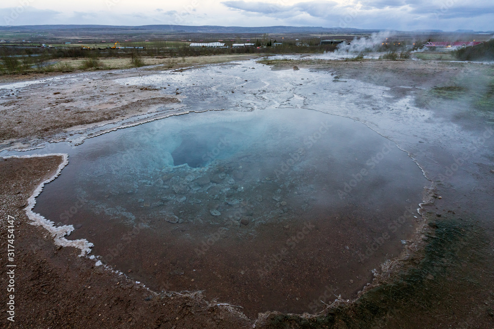 Clear hot pool with big hole towards the ground in the geysir and haukadalur area during blue hour and no people. Smoke from Strokkur and city light in the background. Icelandic traveling concept. Clear hot pool with big hole towards the ground in the geysir and haukadalur area during blue hour and no people. Smoke from Strokkur and city light in the background. Icelandic traveling concept.