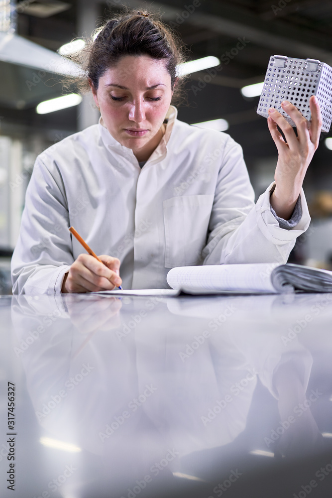 Fototapeta premium Brunette girl in white coat with metal cube sits on table in laboratory