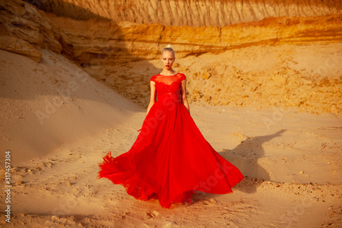 Beauty frontal portrait of a sensual  attractive caucasian young blonde woman  wear in red long flying dress  posing in a sand desert  looking at camera.