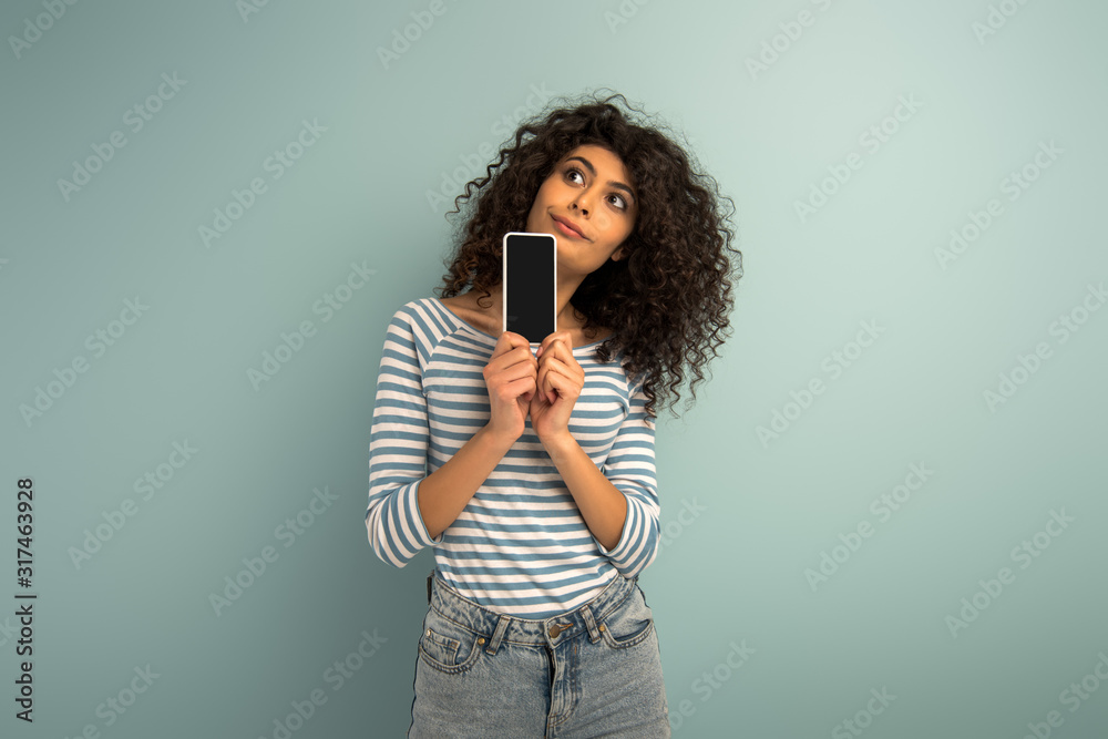 thoughtful mixed race girl looking away while showing smartphone with blank screen on grey background thoughtful mixed race girl looking away while showing smartphone with blank screen on grey background