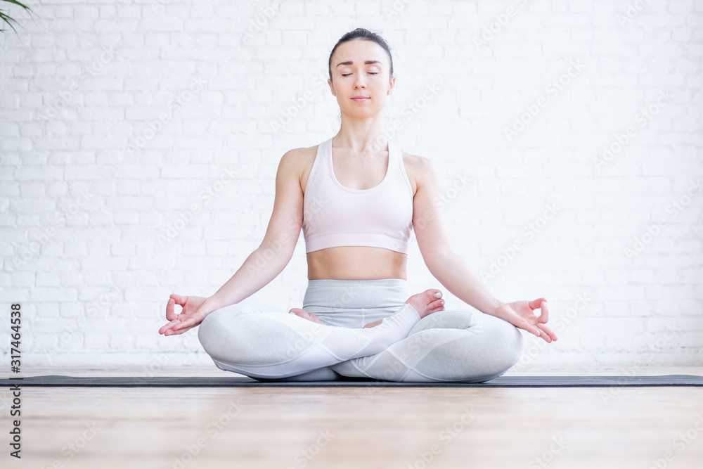 Healthy pacified young woman in tracksuit practices yoga in lotus position and mudra gesture on rug on floor on white brick wall. Healthy respiratory and nervous system concept. Advertising space Healthy pacified young woman in tracksuit practices yoga in lotus position and mudra gesture on rug on floor on white brick wall. Healthy respiratory and nervous system concept. Advertising space