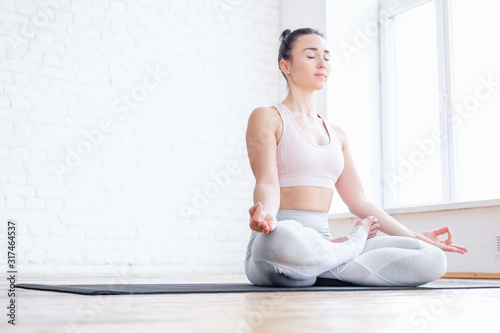 Healthy pacified young woman in tracksuit practices yoga in lotus position and mudra pose on rug on floor on white brick wall. Healthy respiratory and nervous system concept. Advertising space