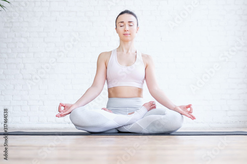 Healthy pacified young woman in tracksuit practices yoga in lotus position and mudra gesture on rug on floor on white brick wall. Healthy respiratory and nervous system concept. Advertising space