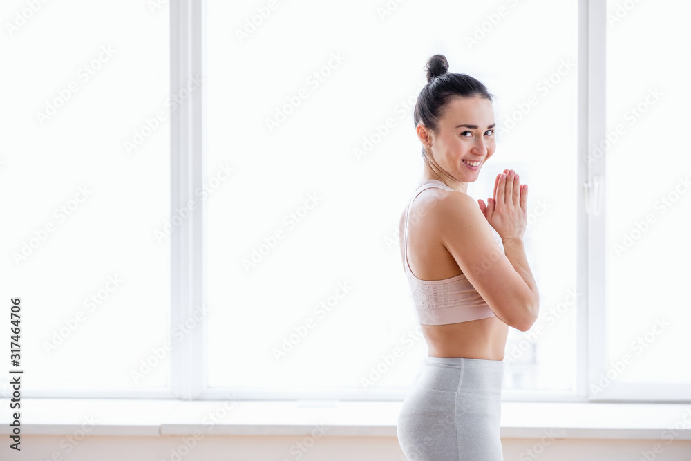 Smiling young woman pilates instructor getting ready for class meditating before exercise on background of large panoramic window. Concept of improving body shape and emotional state. Copyspace