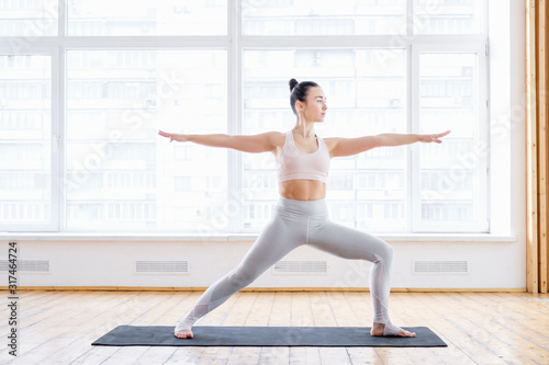 Healthy young beautiful woman doing warrior exercise virabhadrasana pose in tracksuit on rug on floor on background of large window. Concept of basic yoga exercises. Advertising space