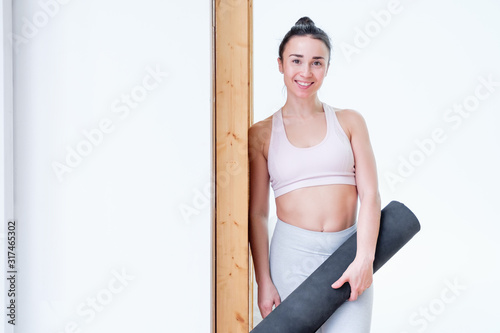 Laughing young positive girl fitness instructor with rug in hands on background of white wall in anticipation of classes. Concept of physical fitness and healthy lifestyle. Advertising space