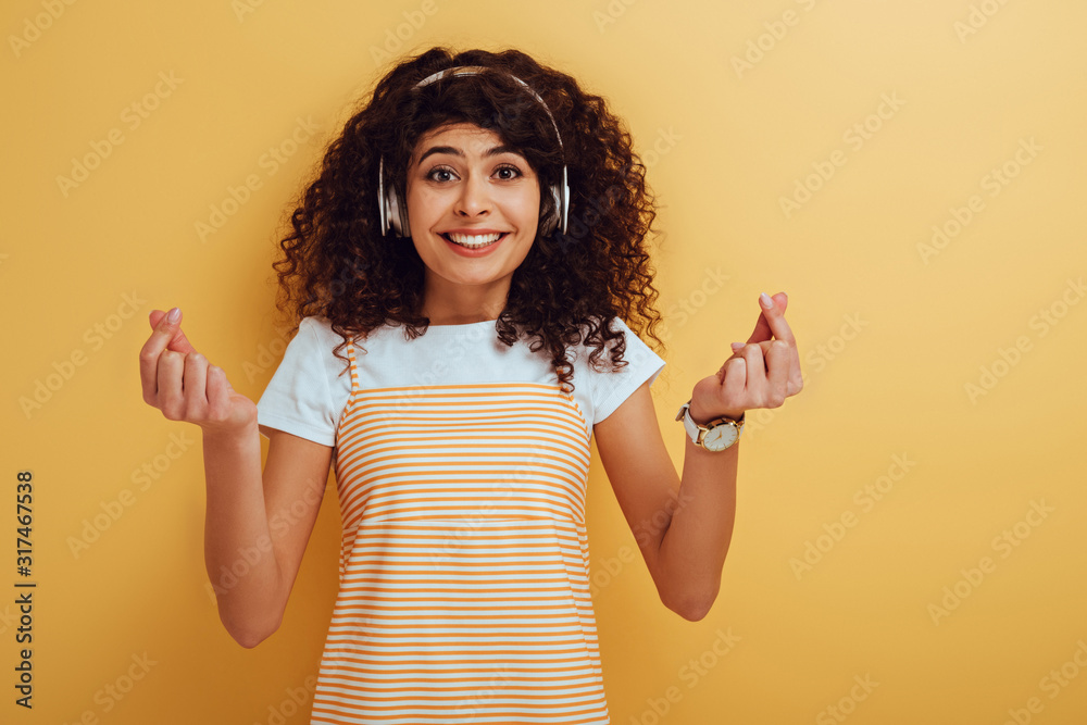 excited mixed race girl in wireless headphones showing winner gesture on yellow background excited mixed race girl in wireless headphones showing winner gesture on yellow background