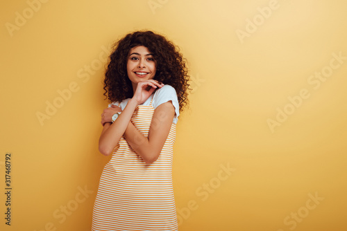 cheerful bi-racial girl holding hand near face while looking at camera on yellow background