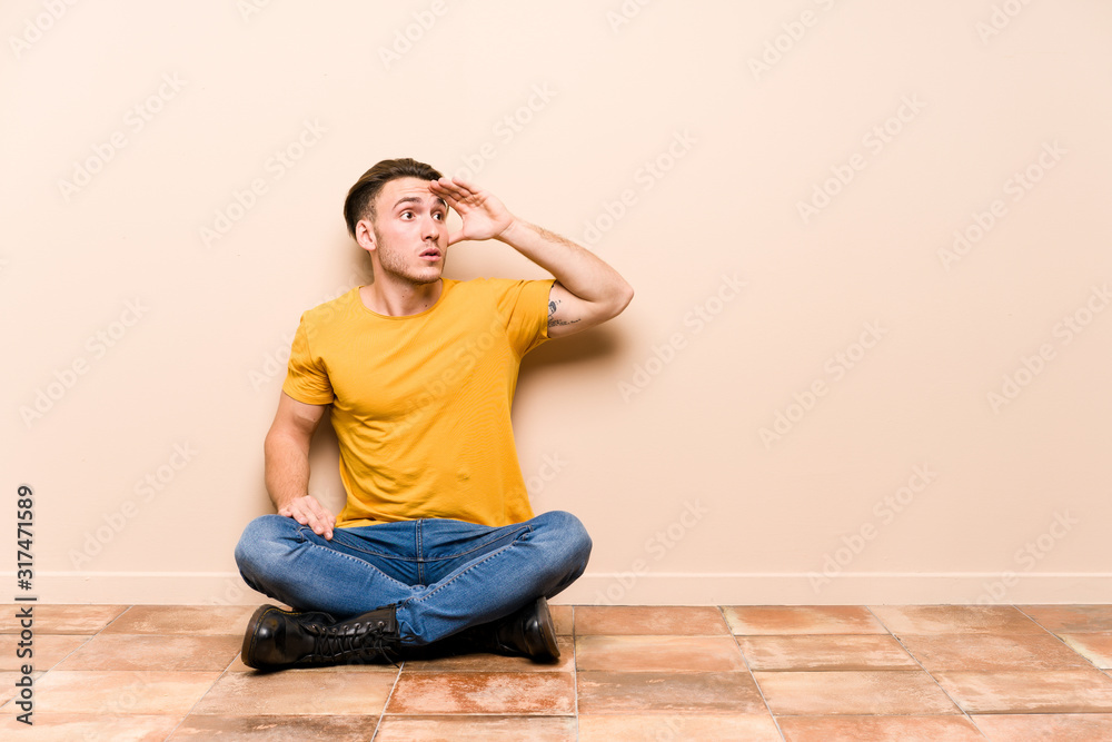 Young caucasian man sitting on the floor isolated looking far away keeping hand on forehead.