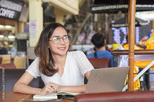 Smiling glasses Asian woman in a white dress sitting in coffee shop, thinking and note to notebook with laptop on table.