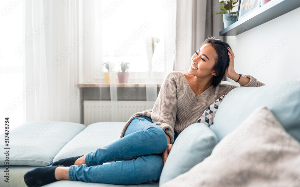 Relaxed smiling asian woman sitting on sofa at home - obrazy, fototapety, plakaty 