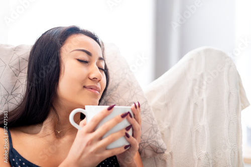 Relaxed young asian woman sitting in armchair with blanket drinking coffee or tea