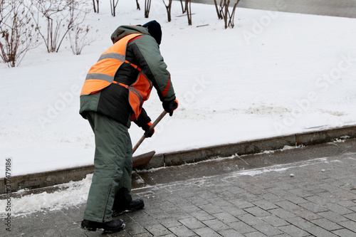 Snow removal in winter, communal services worker in uniform with a shovel clears snow on a street