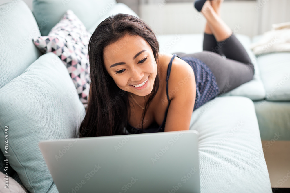 Smiling asian young woman using laptop lying on sofa at home Smiling asian young woman using laptop lying on sofa at home