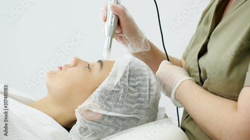 beautician hand moves microdermabrasion device on young woman forehead against white clinic wall close-up