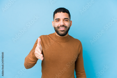 Young latin man against a blue background isolated stretching hand at camera in greeting gesture.