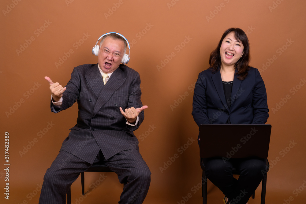 Obraz premium Mature Asian businessman listening to music with mature Asian businesswoman using laptop