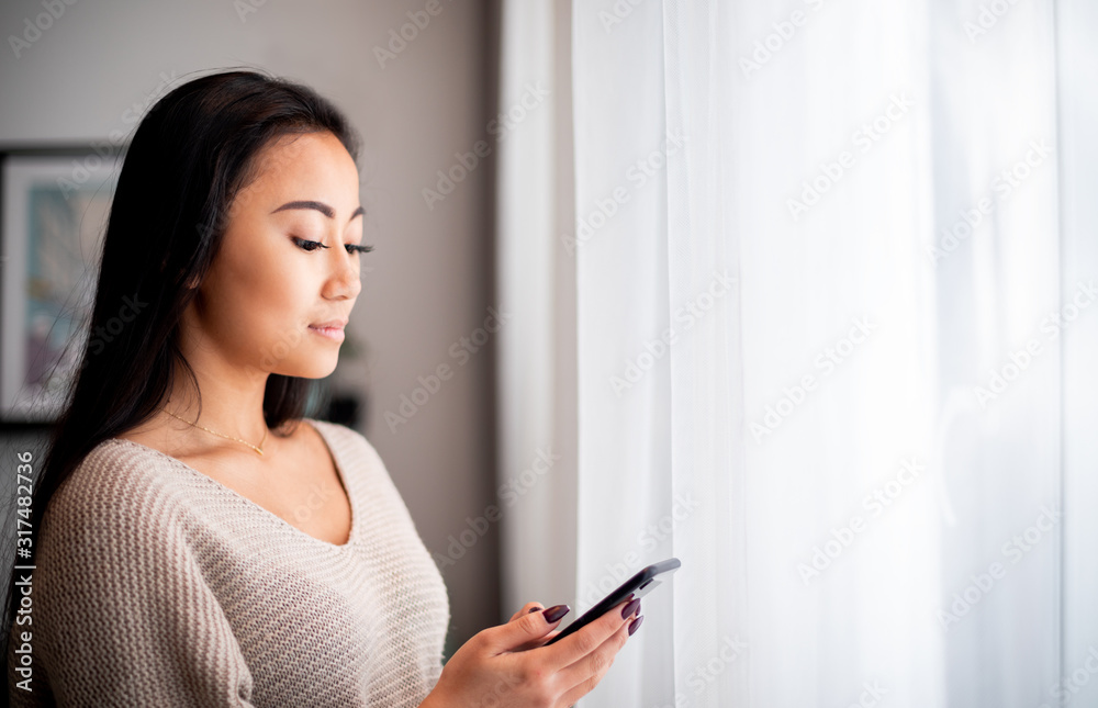 Young asian woman using smartphone at home standing near window Young asian woman using smartphone at home standing near window