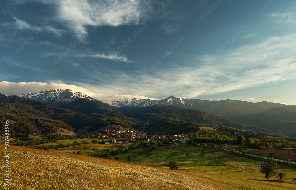 Spring landscape in mountains and blue sky with clouds. Early morning in the mountains, the hamlet against the backdrop of mountains covered with snow. Horizontal orientation. Pirin mountains.