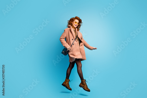 Full body photo of positive cheerful woman jump enjoy beige outerwear vogue style coat travel journey wear cotton pantyhose leather clutch footwear isolated over blue color background