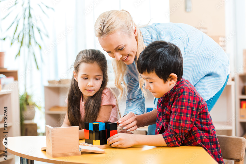 Smiling teacher playing building blocks with kids during lesson in montessori class Smiling teacher playing building blocks with kids during lesson in montessori class