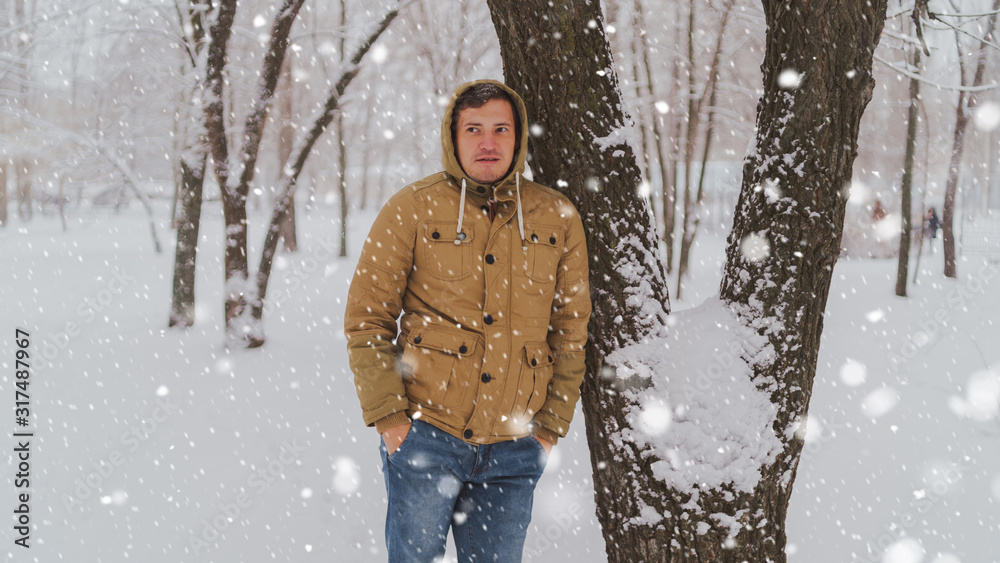 Portrait of young man in jacket and jeans in winter season. Handsome man stands near tree and smiles flirtatiously. Fluffy snow envelops everything around. Portrait of young man in jacket and jeans in winter season. Handsome man stands near tree and smiles flirtatiously. Fluffy snow envelops everything around.