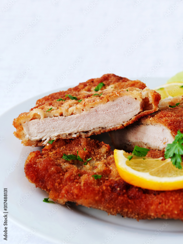 Wiener Schnitzel cut in two served with lemon slices and parsley leaves over white, verical