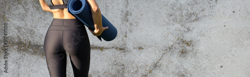 A sporty slim girl in leggings and a top is standing near a concrete wall with a training mat, resting between exercises. photo