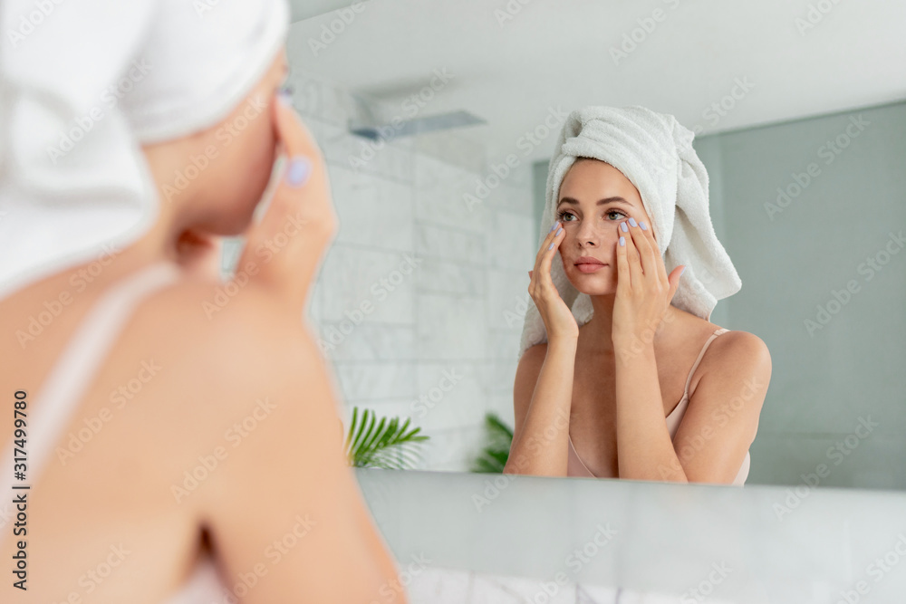 Young woman applying anti-wrinkle eye cream standing behind mirror in home bathroom. Beautiful girl wearing bath towel on head portrait. Cosmetology and beauty procedure. Skin care after cleansing Young woman applying anti-wrinkle eye cream standing behind mirror in home bathroom. Beautiful girl wearing bath towel on head portrait. Cosmetology and beauty procedure. Skin care after cleansing