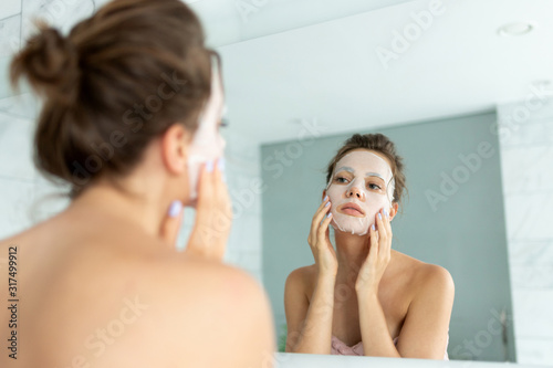 Young woman applying cosmetic mask on face while looking at reflection in mirror.