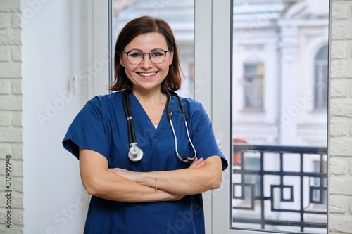 Smiling adult woman doctor in blue uniform stethoscope with folded arms, confident female medic looking at camera, standing near the window in clinic