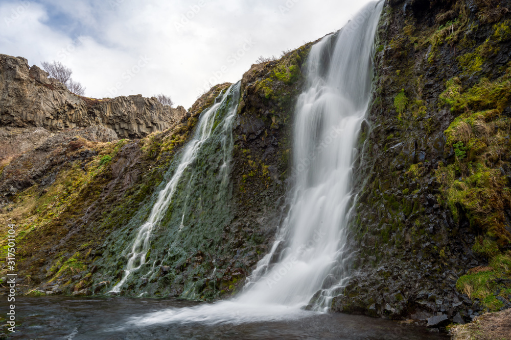 Waterfall cascade in the area of Gjain in Porsdalur in the Icelandic highlands. Overcast sky. Long exposure shot of the waterfall. Green textured rock formation.