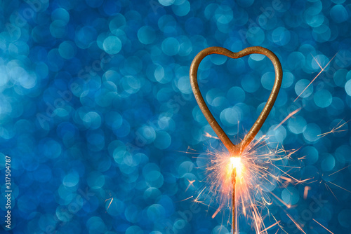 Burning sparkler in heart love shape over blue festive glitter background. Wedding or valentine's day banner with copy-space.
