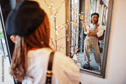 African american woman in overalls and beret against mirror in soft light room.